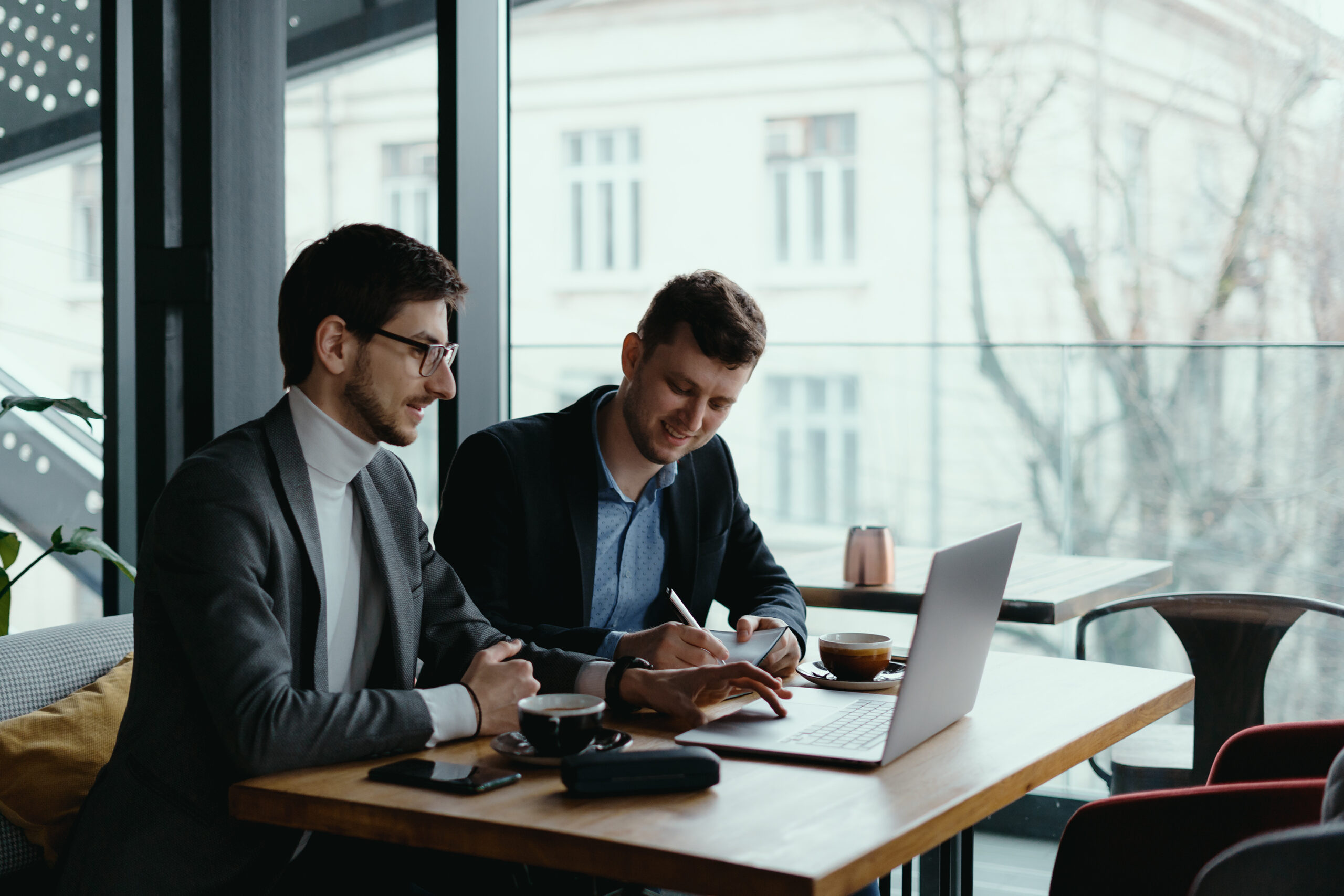Two businessmen talking about Manifest business consulation for Dubai Company Formation sitting with laptop at desk near large window, planning project, considering business offer, sharing ideas while drinking coffee together